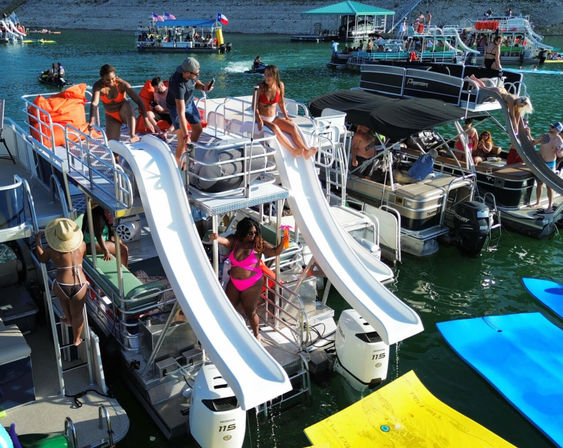 Summer lake party with pontoon boats fitted with twin white waterslides, groups in swimsuits sliding and lounging by green water around colorful floating mats.