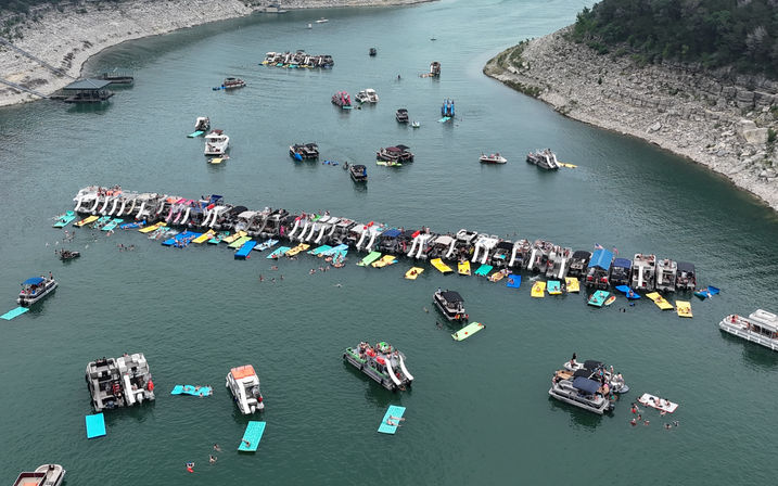 Aerial view of a lively lake cove with dozens of boats and pontoons forming a floating party line, colorful inflatable mats and slides, swimmers in green-blue water and rocky shoreline