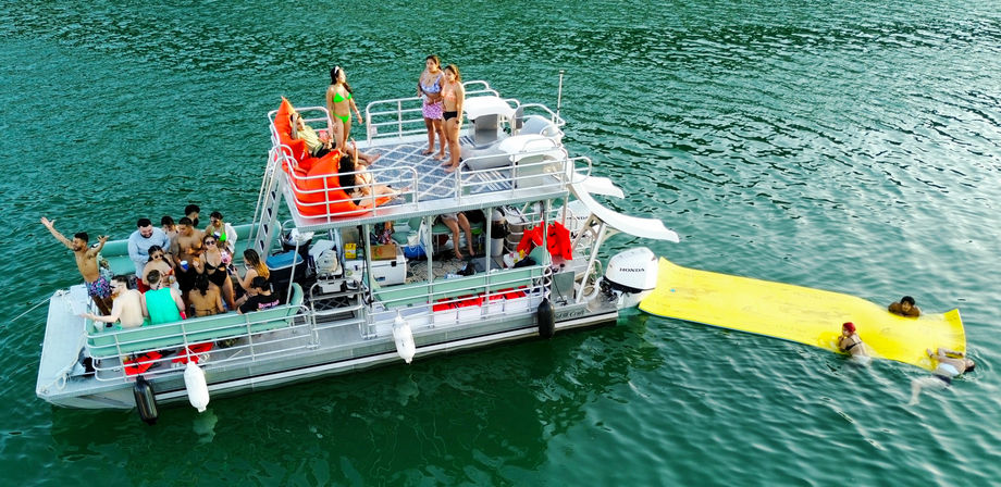 Aerial view of a summer pontoon boat party on a green lake, friends socializing on a two-level deck with orange bean bags and a large yellow floating mat with swimmers relaxing in the water.