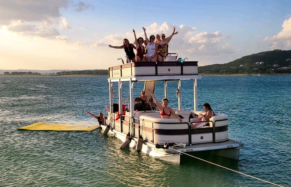 Group of friends cheering on a double-decker pontoon boat anchored in a sunlit bay at sunset, with people lounging onboard and a swimmer on a yellow floating mat nearby.