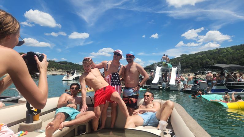 Five friends posing on a pontoon boat at a busy summer lake party, photographer at left, inflatable water slides and other boats on the water under a bright blue sky