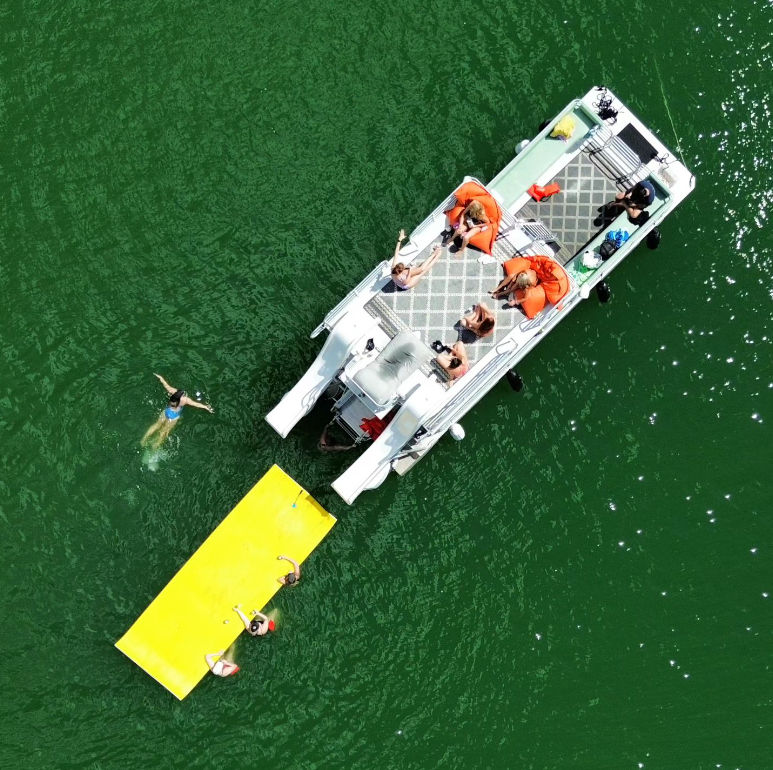 Aerial drone view of a pontoon boat on a green lake with people lounging on deck in orange float chairs and swimmers playing on a bright yellow floating mat