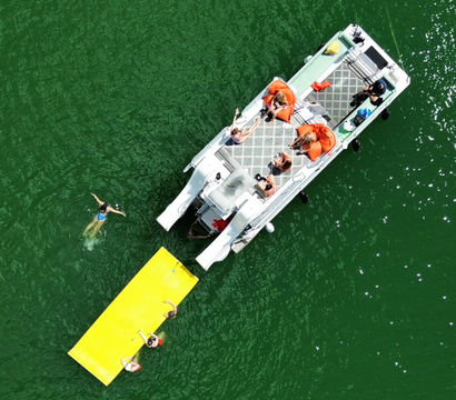 Aerial drone view of a pontoon boat on a green lake with people lounging on deck in orange float chairs and swimmers playing on a bright yellow floating mat