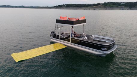 Black pontoon boat with upper deck lounge and bright yellow floating mat drifting on a calm lake near a tree-lined shoreline, two people relaxing onboard.