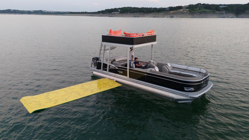 Black pontoon boat with upper deck lounge and bright yellow floating mat drifting on a calm lake near a tree-lined shoreline, two people relaxing onboard.