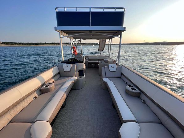 Pontoon boat interior with plush cushioned lounge seating, built‑in cup holders and upper-deck canopy cruising on calm lake waters at sunset