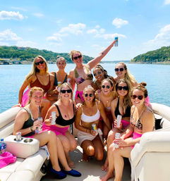Summer pontoon boat party on a sunny lake — a smiling group of friends in swimsuits and sunglasses holding canned drinks, with tree-lined shoreline and blue sky in the background.