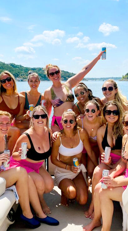 Summer pontoon boat party on a sunny lake — a smiling group of friends in swimsuits and sunglasses holding canned drinks, with tree-lined shoreline and blue sky in the background.
