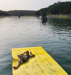 Person in a swimsuit lounging on a bright yellow floating mat on a calm lake, making peace signs while boats drift near a tree-covered shoreline.
