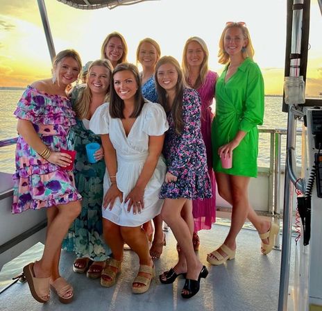 Eight women in colorful summer dresses smiling and posing on a boat at sunset over calm water, holding drinks on a sunset cruise