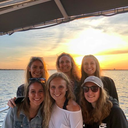 Six friends smiling together on a boat during golden-hour sunset, calm water and a glowing sky in the background.