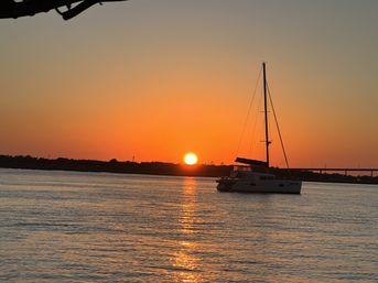 Sailboat anchored in a calm bay at golden sunset, sun dipping behind a low shoreline and bridge and casting a shimmering reflection across the water.