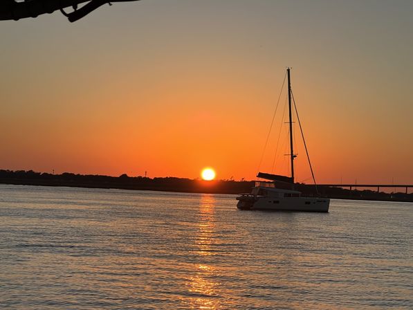 Sailboat anchored in a calm bay at golden sunset, sun dipping behind a low shoreline and bridge and casting a shimmering reflection across the water.