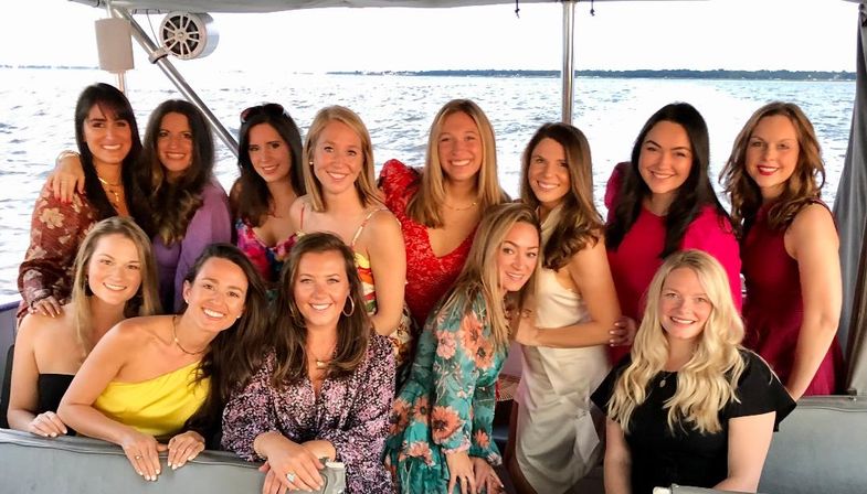 Smiling group of friends in colorful summer dresses posing on a boat over a calm lake with distant shoreline and warm sunset light