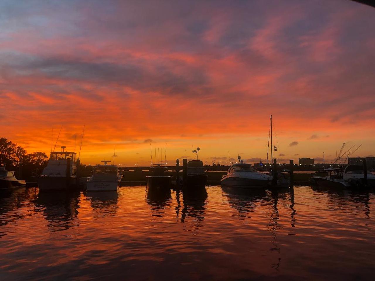 Vibrant orange and pink sunset over a coastal marina, silhouetted boats and masts reflected in calm water