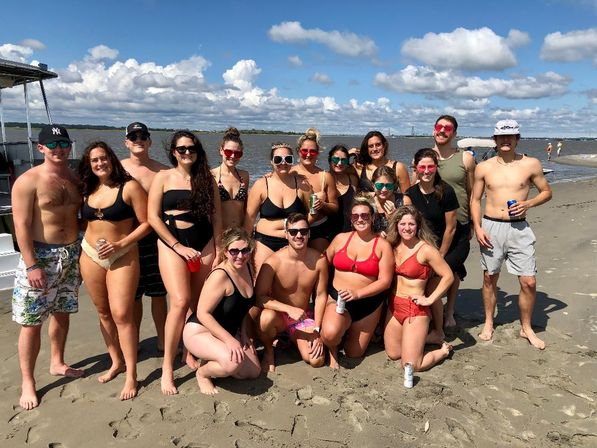 Large group of friends in colorful swimsuits posing on a sandy coastal beach by a calm bay under a bright blue sky with puffy clouds