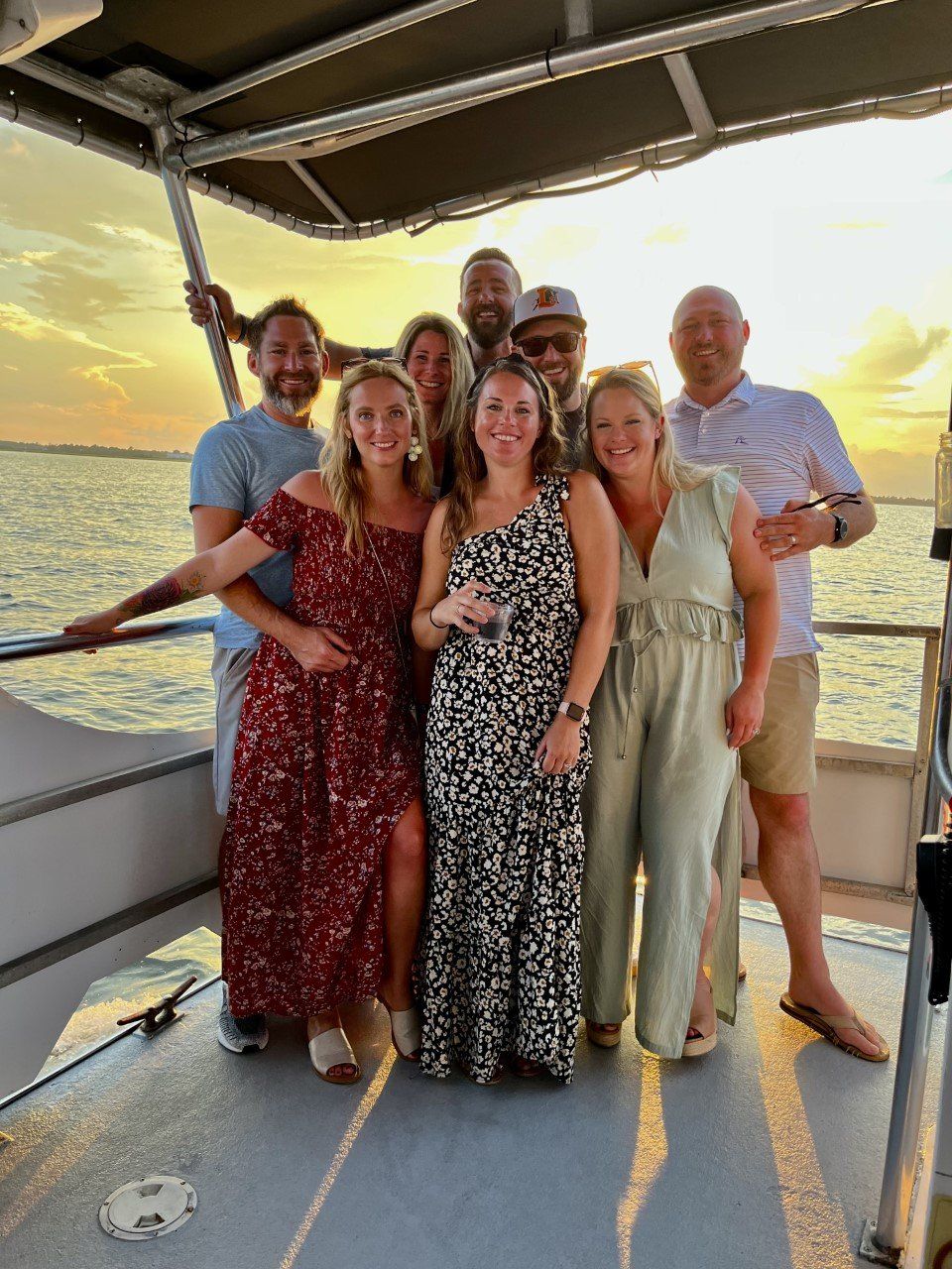 Group of eight people smiling on a boat deck during a golden sunset cruise over calm water, wearing summer dresses and casual shorts — waterfront evening vibe.
