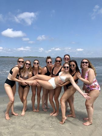 Nine women in swimsuits on a sunny sandy beach by calm coastal waters, playfully holding one woman horizontally under a blue sky with scattered clouds.