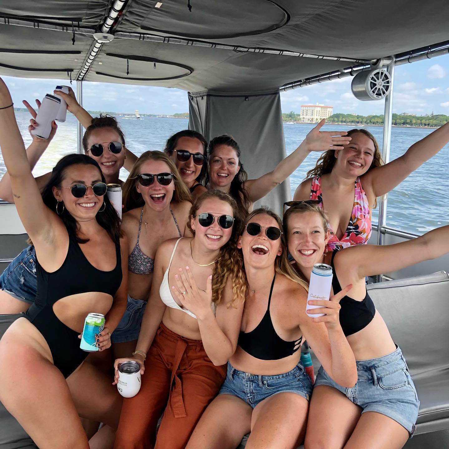 Group of nine women in swimsuits and sunglasses cheering and holding drinks on a covered pontoon boat during a sunny summer boat party, with calm bay waters and a distant waterfront building in the background.
