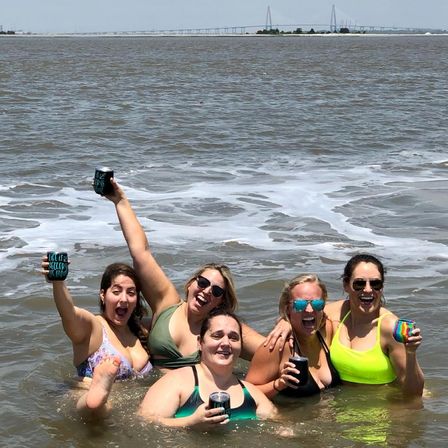 Five friends in swimsuits waist-deep in coastal water, laughing and raising canned drinks amid foamy surf with a distant suspension bridge on the horizon on a sunny beach day
