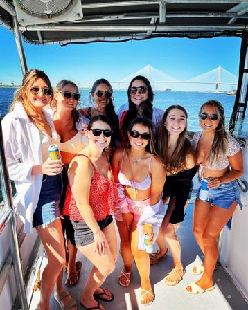 Smiling group of young women in swimsuits and sunglasses posing on a sunny boat deck, holding drinks with blue harbor water and a cable-stayed bridge in the background.