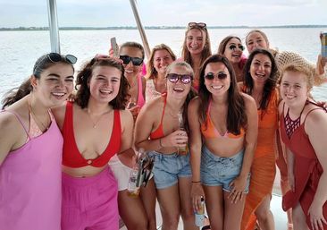 Group of smiling women on a boat at a lake, wearing swimsuits and summer dresses and holding drinks during a sunny boat party.