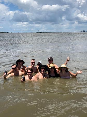 Group of people waist-deep in brown coastal water, smiling and cheering with canned drinks under a dramatic cloudy sky, with a distant suspension bridge and cargo ship on the horizon.