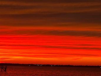 Vivid red-orange sunset with layered clouds over a calm waterfront, silhouetted pier at left and distant shoreline lights.