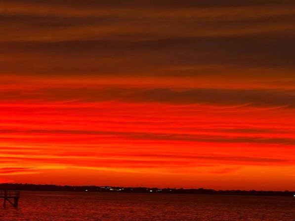 Vivid red-orange sunset with layered clouds over a calm waterfront, silhouetted pier at left and distant shoreline lights.