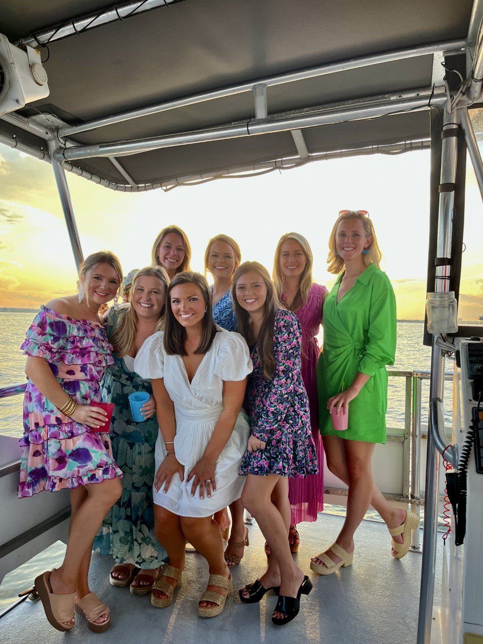 Eight women in colorful summer dresses smiling together on a boat deck during a golden sunset over calm coastal waters, some holding cups.