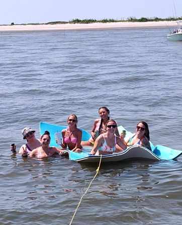 Six friends in bikinis laughing and holding drinks while floating on a blue foam mat in calm coastal water near a sandy beach and anchored boat — summer beach outing