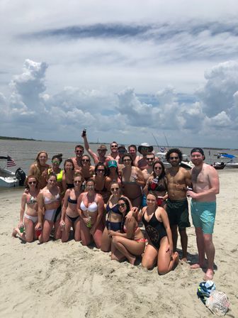 Large group of friends in colorful swimsuits posing on a sandy coastal beach by anchored boats and a calm bay, holding drinks under a dramatic partly cloudy summer sky.