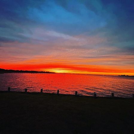 Vibrant red-orange sunset over a coastal bay at dusk, silhouetted shoreline and pier posts reflecting on rippling water.