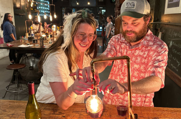 Smiling man and woman adjust a glowing Edison bulb over a wooden bar table with wine and warm industrial pendant lights in a cozy urban craft bar at night