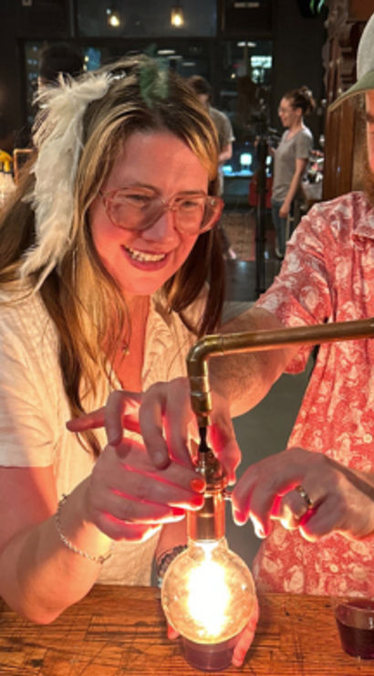 Smiling man and woman adjust a glowing Edison bulb over a wooden bar table with wine and warm industrial pendant lights in a cozy urban craft bar at night