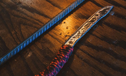 Rusty handmade metal blade with red-and-black paracord-wrapped handle beside a textured rebar on a weathered wooden workbench lit by warm workshop light