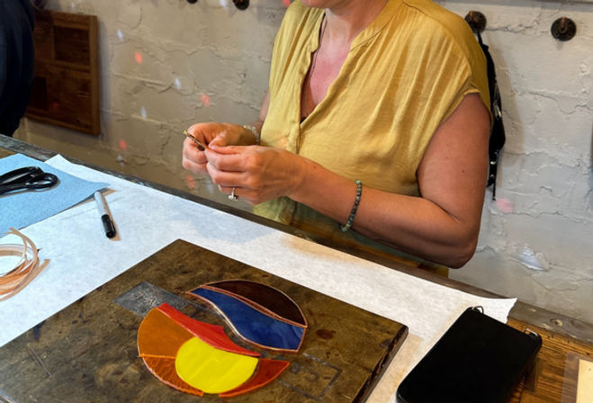 Hands of a person in a yellow top crafting colorful stained-glass pieces on a workshop table with scissors, marker, and a smartphone nearby