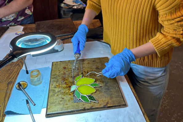 Person in a yellow sweater wearing blue gloves soldering a green stained-glass leaf on a metal workboard under a magnifying lamp in a craft studio, with flux jar and tools nearby.