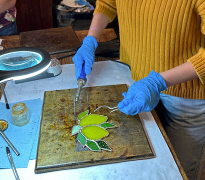 Person in a yellow sweater wearing blue gloves soldering a green stained-glass leaf on a metal workboard under a magnifying lamp in a craft studio, with flux jar and tools nearby.