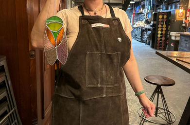 Artist in a brown leather apron holding a colorful stained-glass suncatcher (green, orange, purple panels) inside a busy craft workshop with stools and shelving
