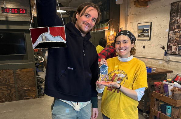 Two smiling crafters in a rustic stained-glass workshop holding colorful handmade stained-glass pieces — DIY art studio scene