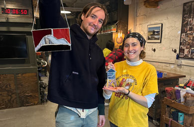 Two smiling crafters in a rustic stained-glass workshop holding colorful handmade stained-glass pieces — DIY art studio scene
