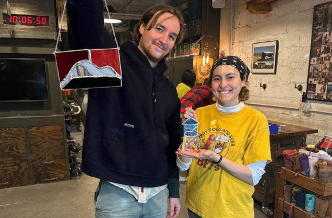 Two smiling crafters in a rustic stained-glass workshop holding colorful handmade stained-glass pieces — DIY art studio scene