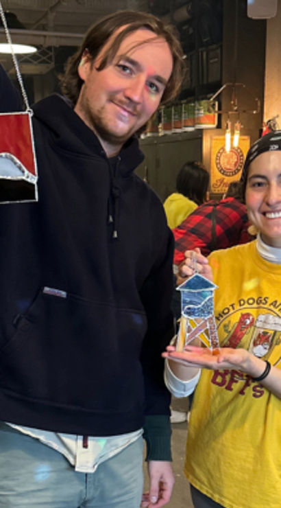 Two smiling crafters in a rustic stained-glass workshop holding colorful handmade stained-glass pieces — DIY art studio scene