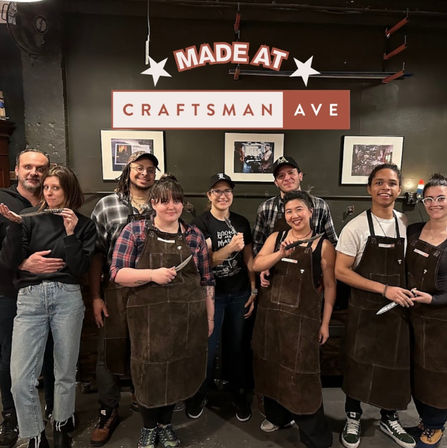 Smiling group of students in leather aprons holding handmade knives in a hands-on knife-making craft studio workshop