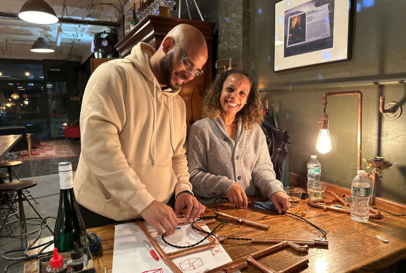 Two smiling people assembling copper-pipe frames on a wooden counter with sketches, tools, water bottles and a warm exposed-bulb lamp in a cozy indoor workshop setting.