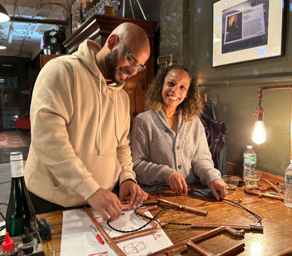Two smiling people assembling copper-pipe frames on a wooden counter with sketches, tools, water bottles and a warm exposed-bulb lamp in a cozy indoor workshop setting.