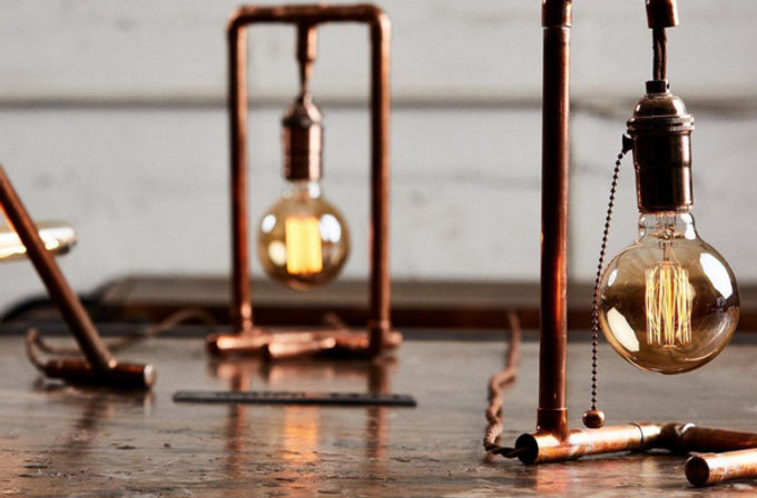 Rustic industrial scene: glowing Edison bulbs hanging from copper-pipe table lamps on a worn wooden tabletop