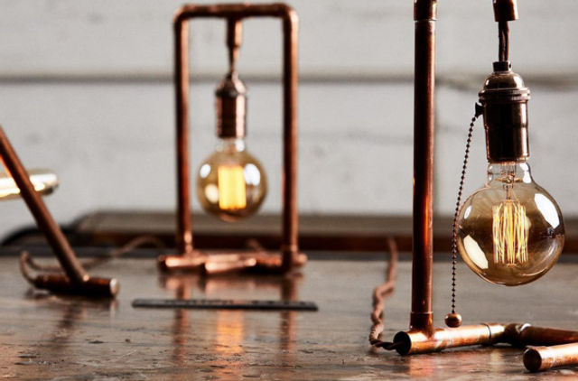 Rustic industrial scene: glowing Edison bulbs hanging from copper-pipe table lamps on a worn wooden tabletop