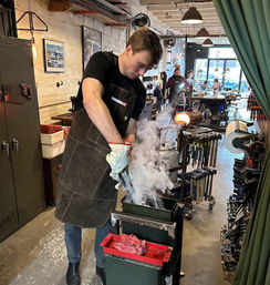 Artisan wearing apron and gloves dunking glowing molten glass into a water pan, steam rising in a busy glassblowing studio with furnace, tools, and visitors in the background.